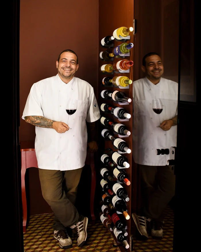 Chef Willian Vieira sorrindo, de dólmã branco e taça de vinho, ao lado da adega do La Faet
